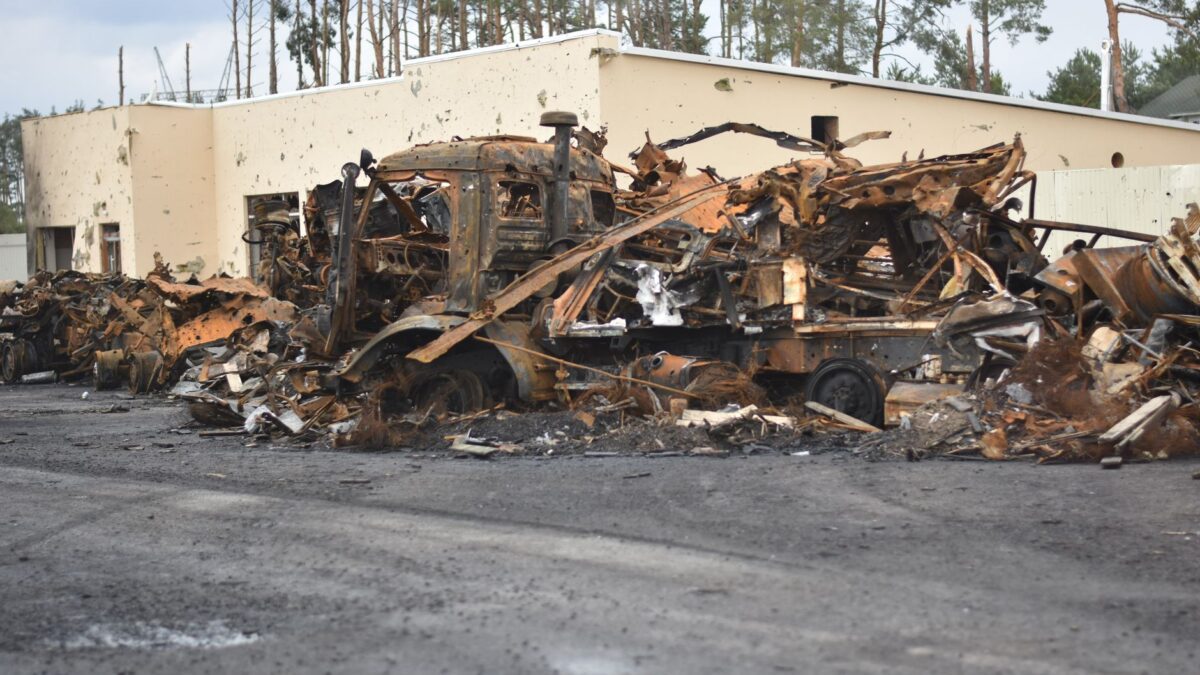 Burned vehicles and building destruction create a war-torn scene outdoors.