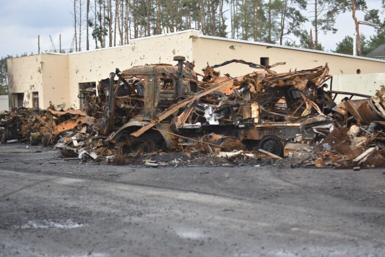 Burned vehicles and building destruction create a war-torn scene outdoors.